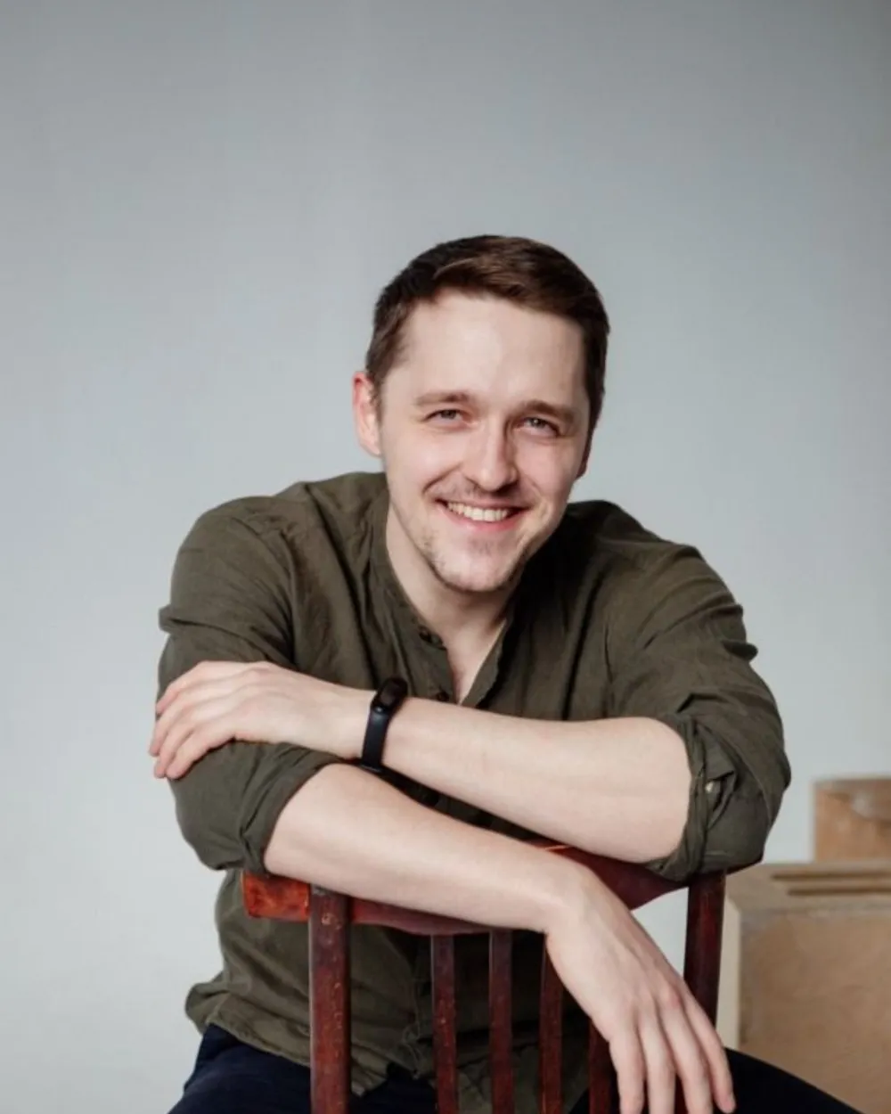 Vasilii smiling, sitting backwards on a wooden chair in an olive shirt.